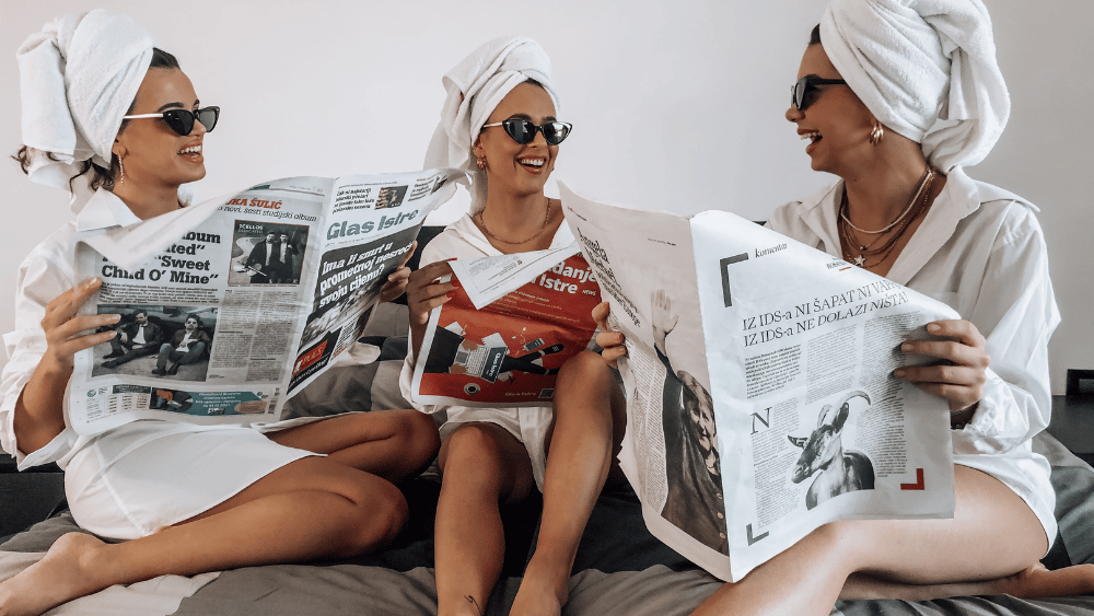 3 Gen Z friends sitting together in hair towels reading newspapers - Dig & Dig